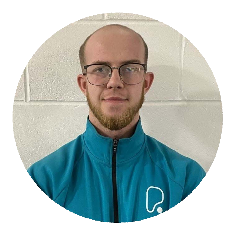 A man with short hair, beard, and glasses wearing a teal zip-up jacket against a white wall.