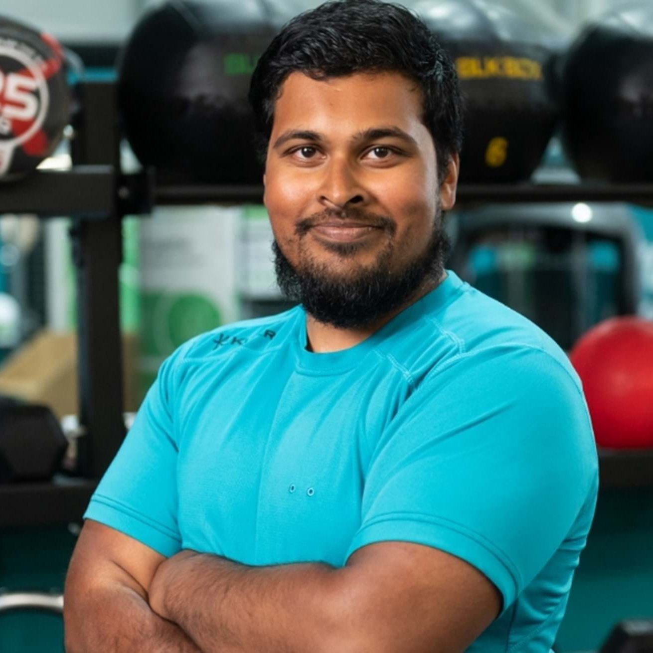 A smiling man with a beard wearing a blue athletic shirt, standing in front of a rack of gym balls.