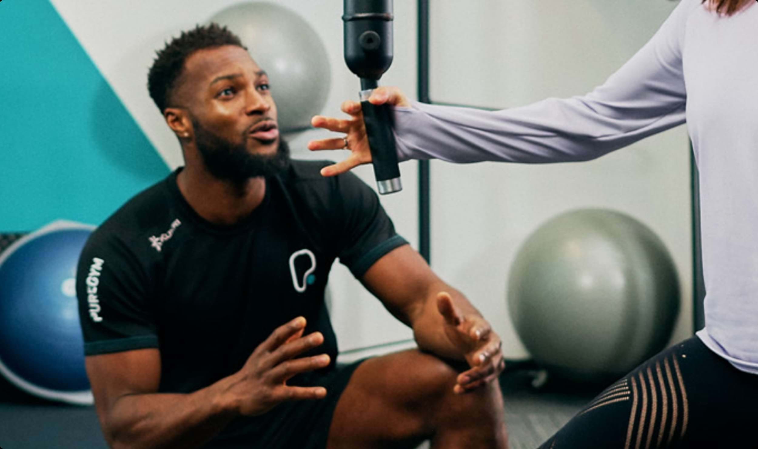 A PureGym personal trainer instructing a client using a massage gun in a gym setting.