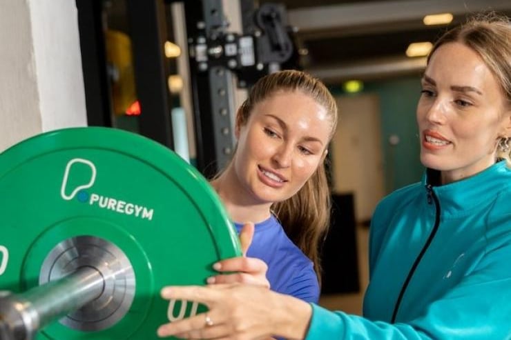 A female gym instructor in a blue jacket guiding a client on using a PureGym weight plate.