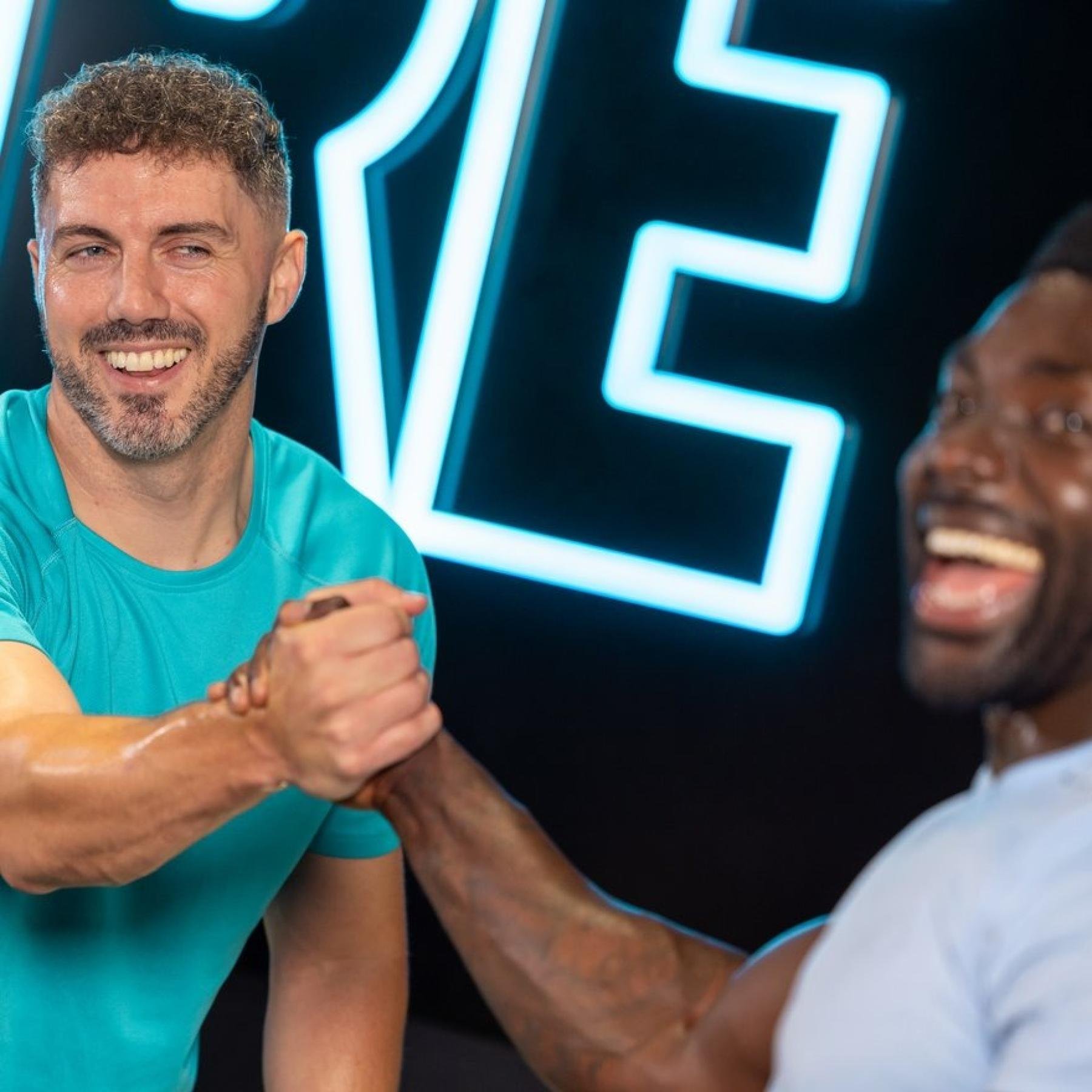 Two men shaking hands in a gym, smiling with neon lighting in the background.