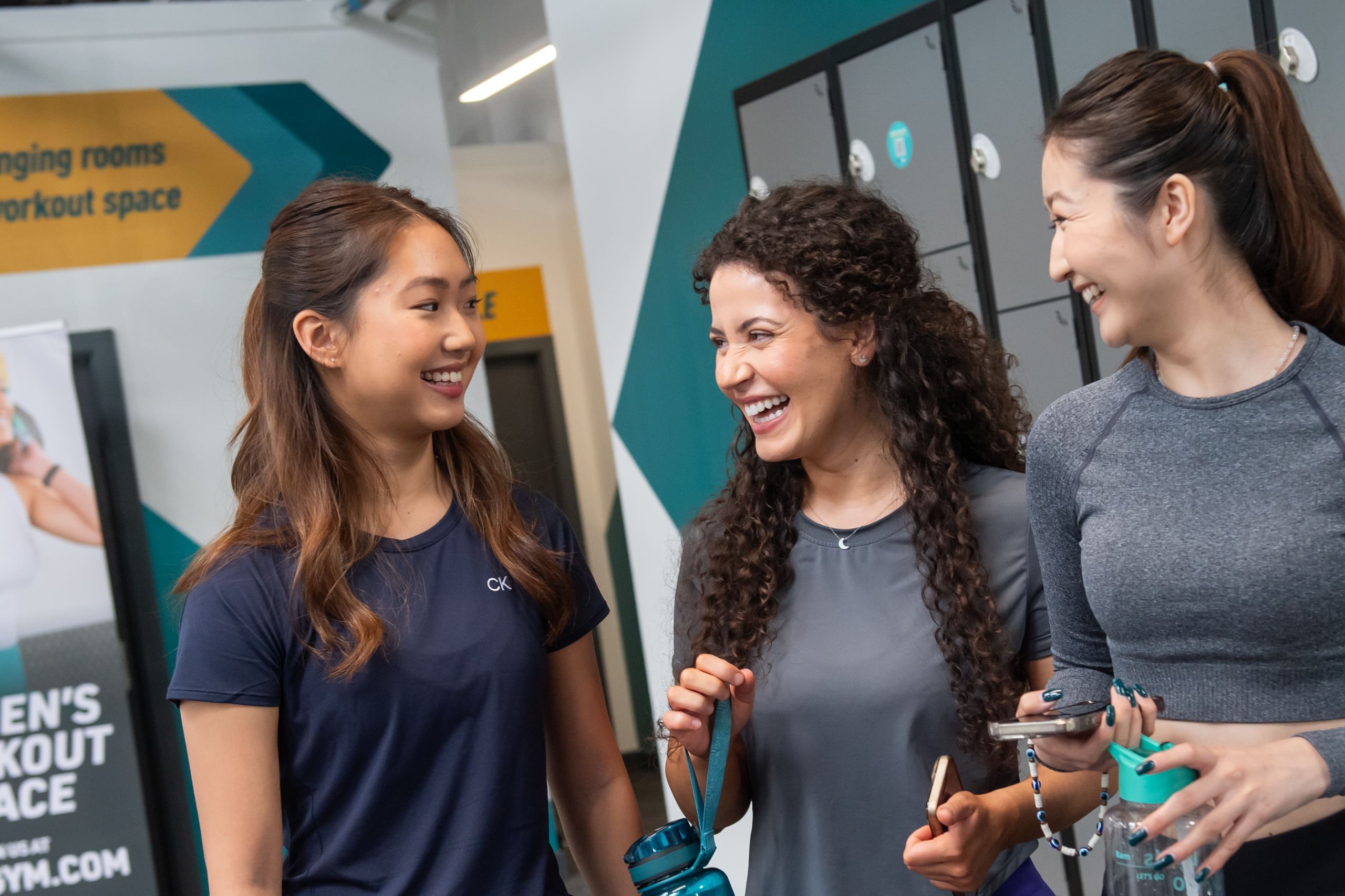 Three young women laughing and talking together near lockers in a modern gym.