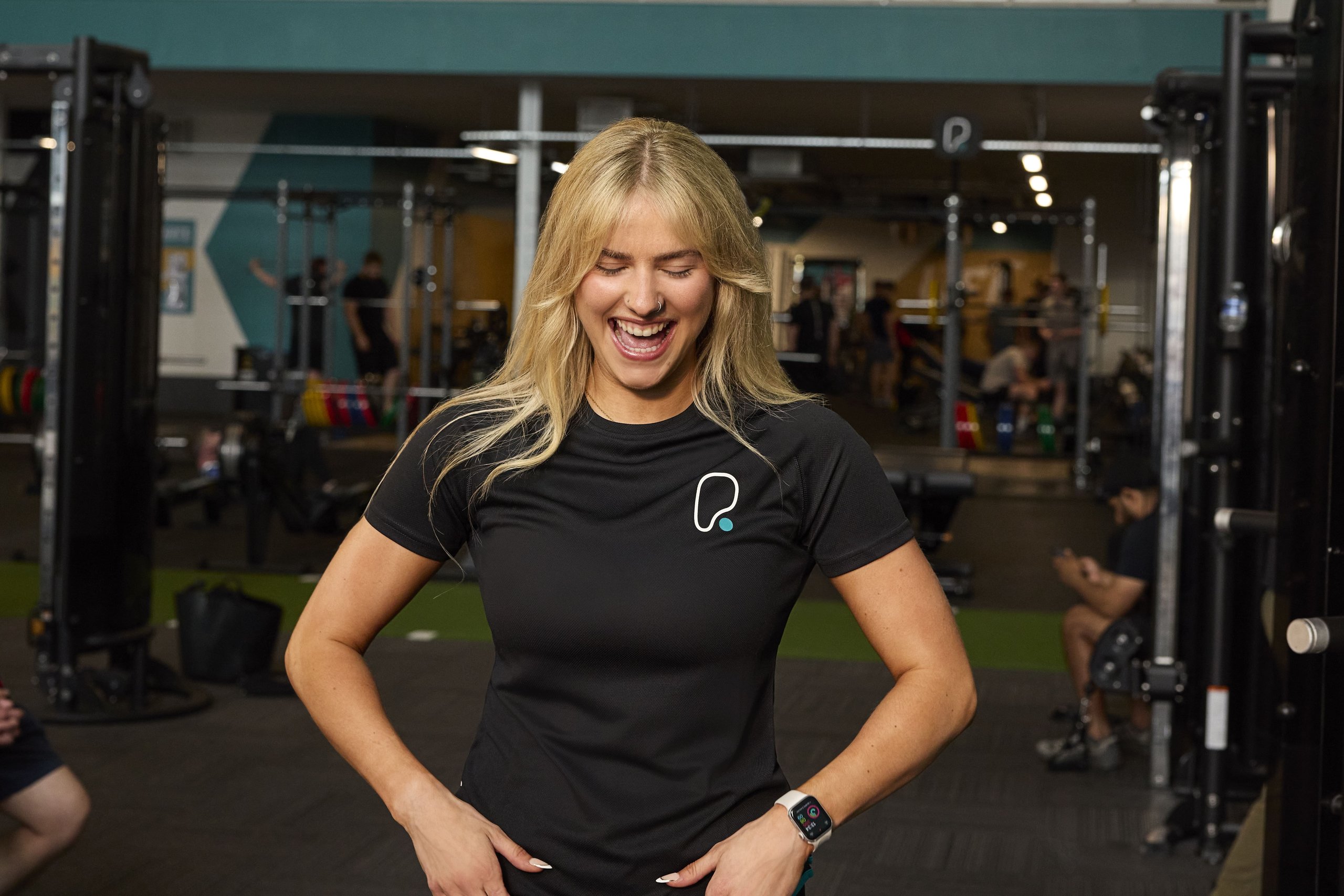 A smiling blonde woman in a black t-shirt laughs inside a well-equipped gym with other people.