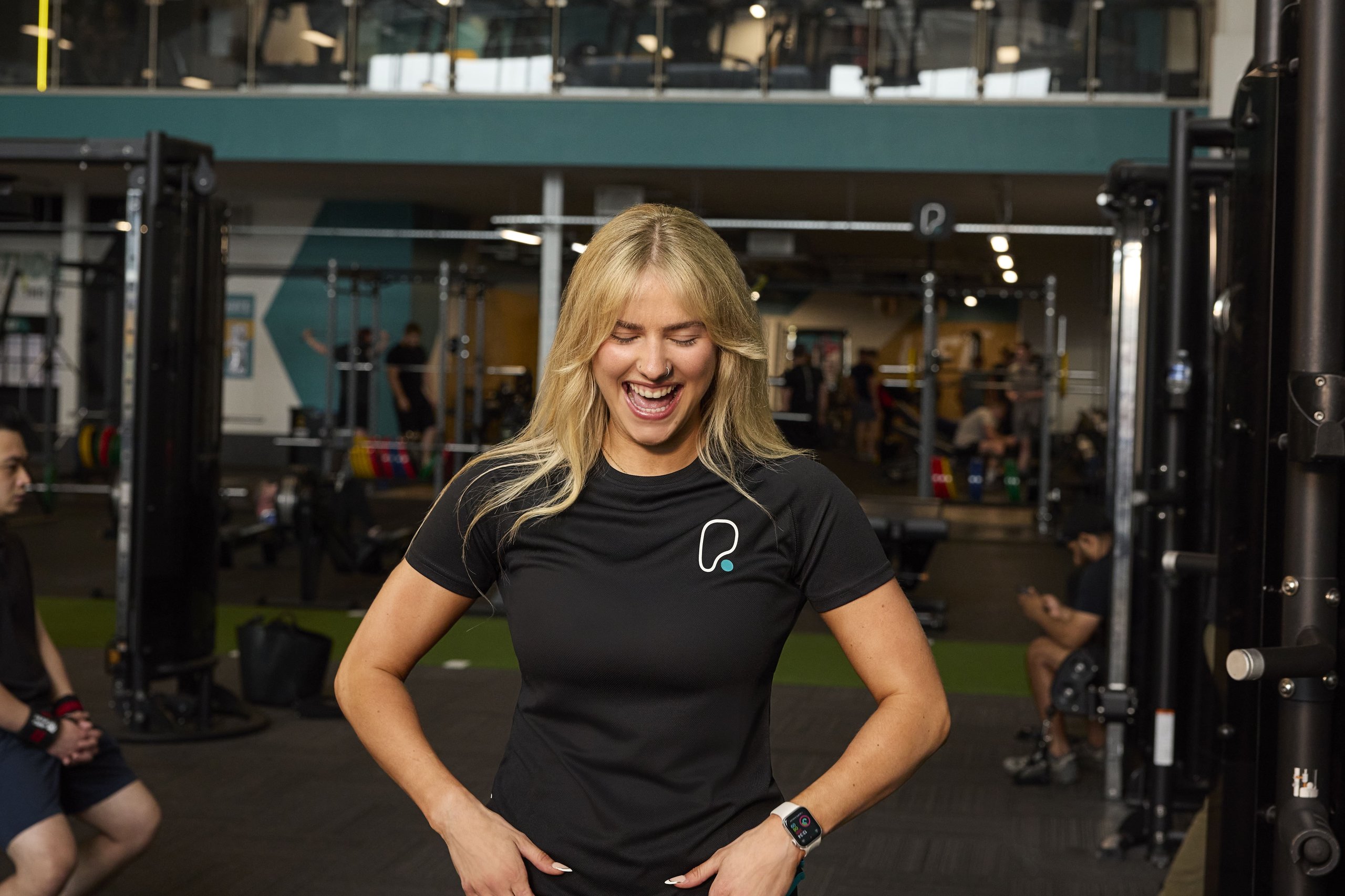 A smiling blonde woman in a black t-shirt laughs inside a well-equipped gym with other people.