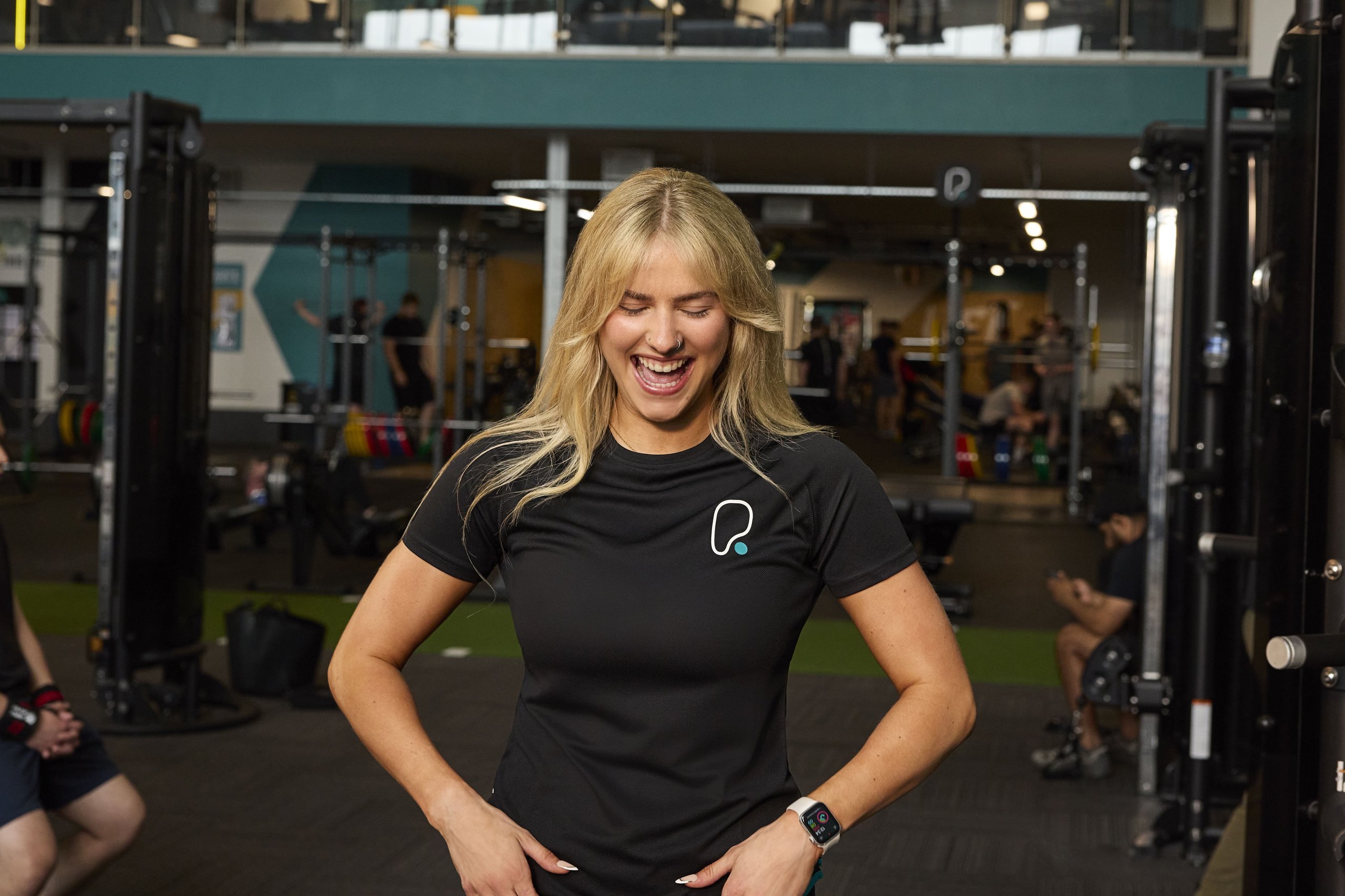 A smiling blonde woman in a black t-shirt laughs inside a well-equipped gym with other people.