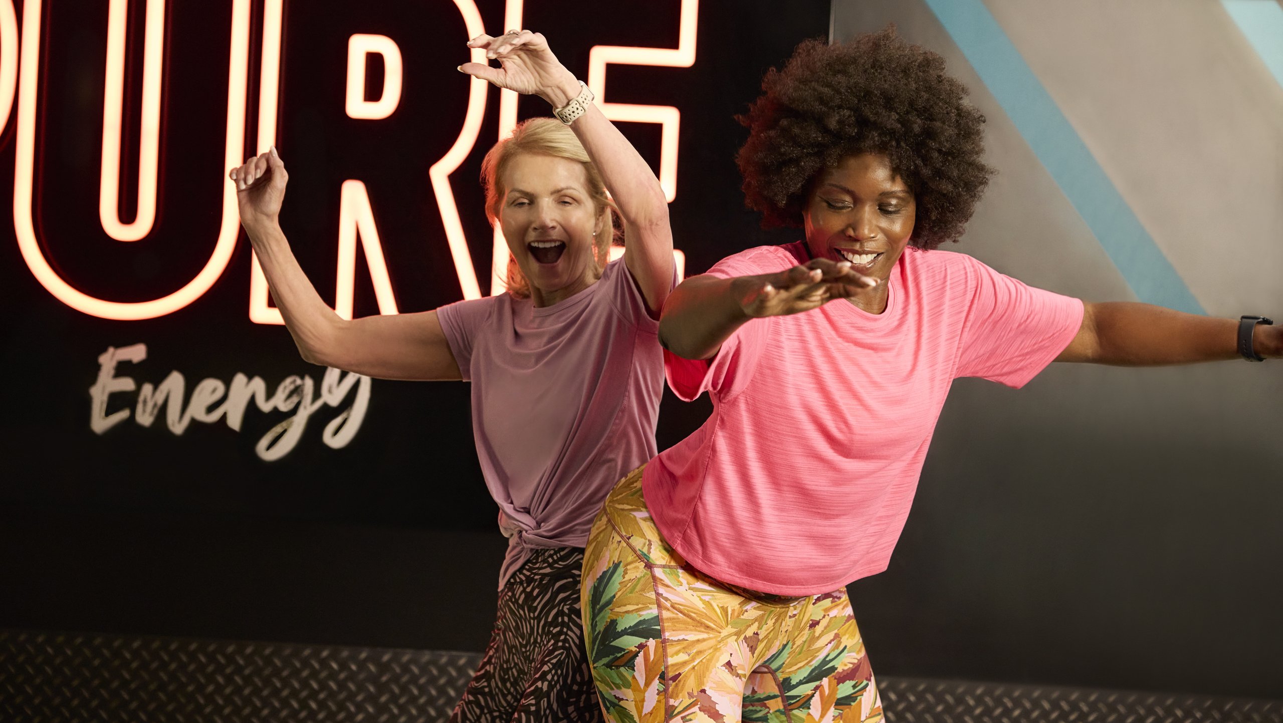 Two women happily dancing together in front of a glowing sign in a gym.