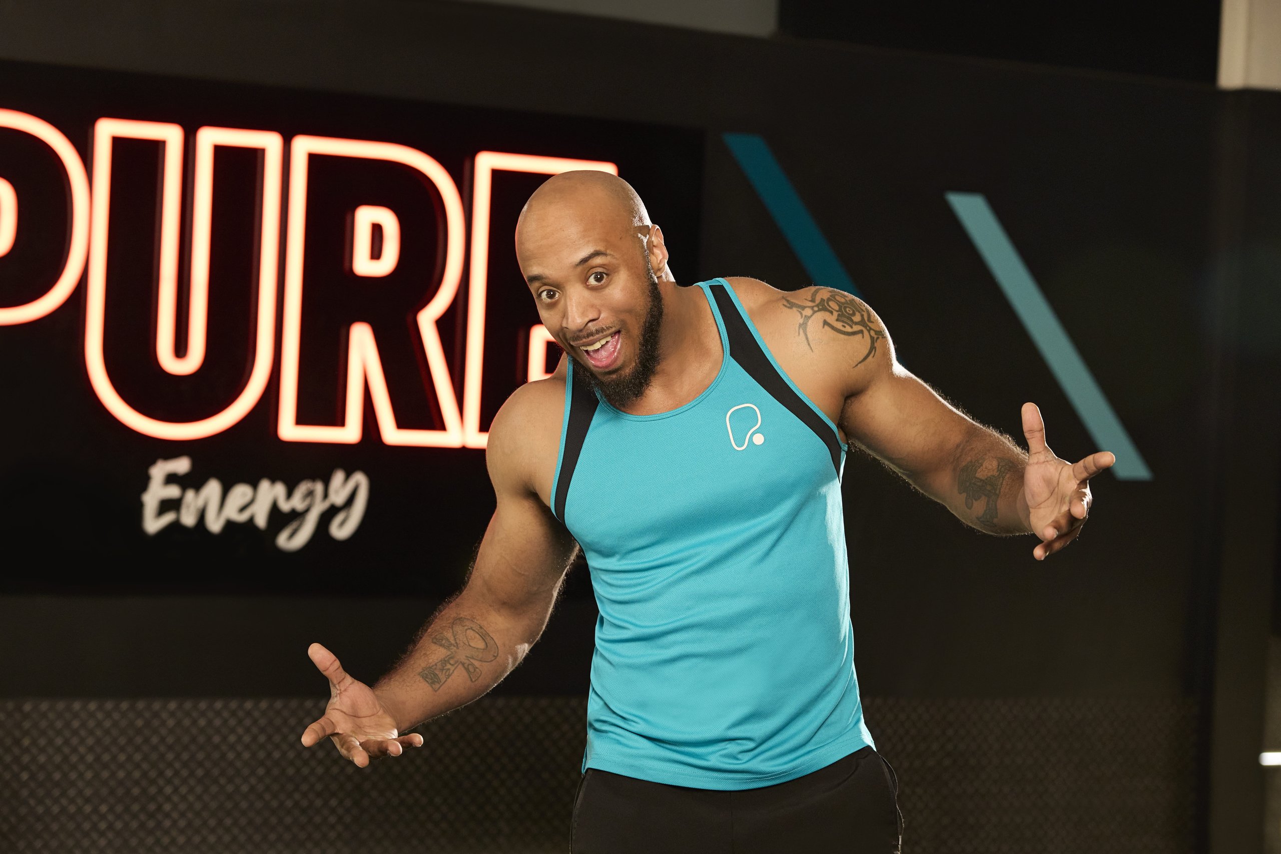 A smiling bald man in a blue tank top stands in a gym with a neon sign in the background.