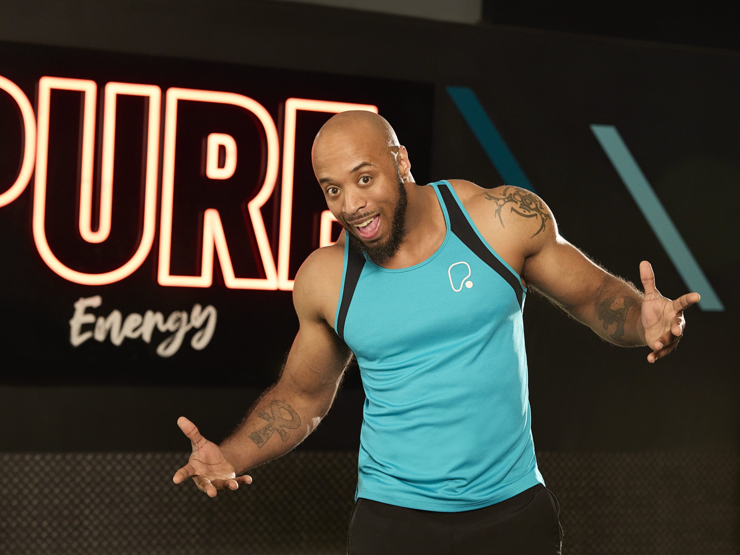 A smiling bald man in a blue tank top stands in a gym with a neon sign in the background.