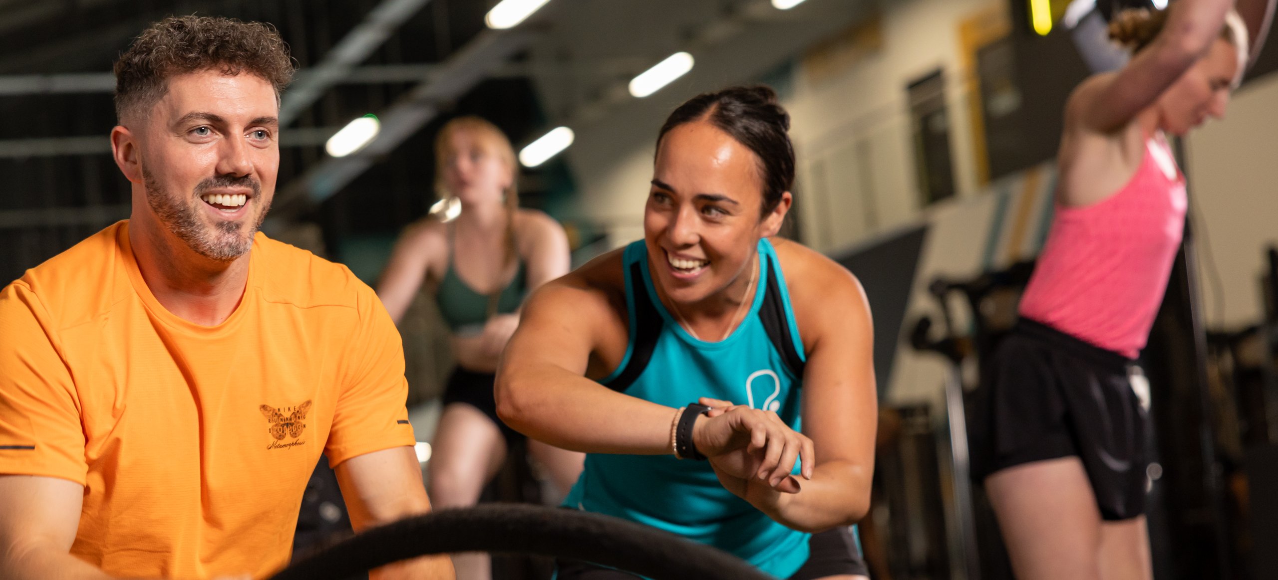 A man smiling while using battle ropes in a gym, with a woman looking at her watch nearby.